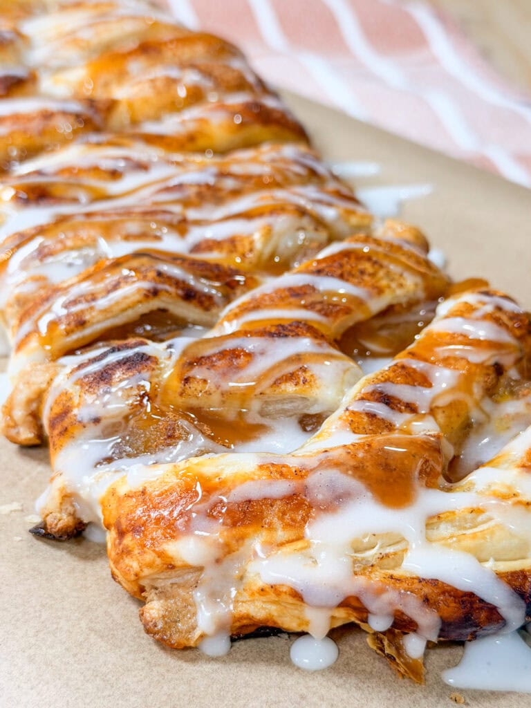 A close-up of a braided pastry topped with white icing and caramel sauce, resting on parchment paper with a striped cloth in the background.