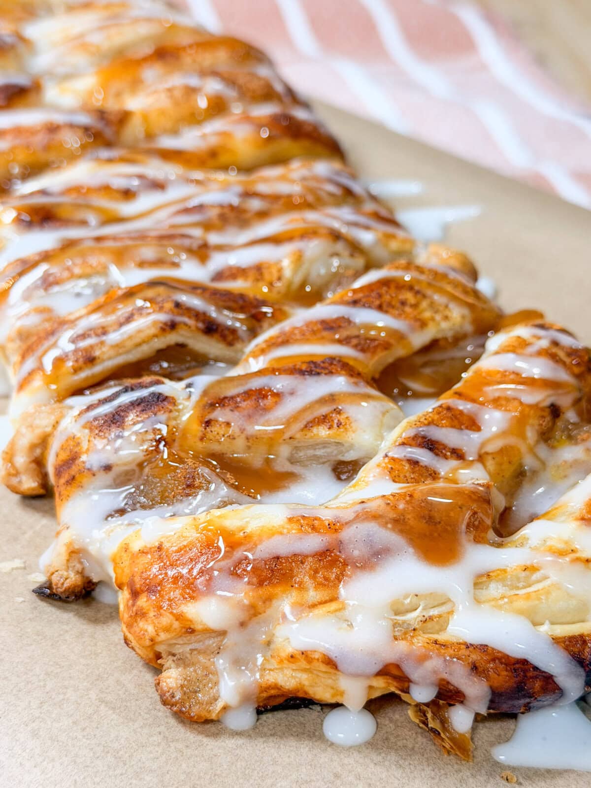 A close-up of a braided pastry topped with white icing and caramel sauce, resting on parchment paper with a striped cloth in the background.