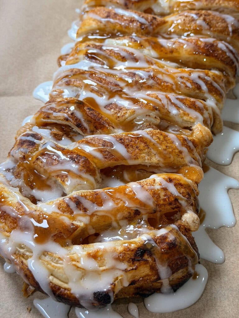 A close-up of a braided pastry with golden, flaky layers, topped with white icing drizzle, resting on parchment paper.