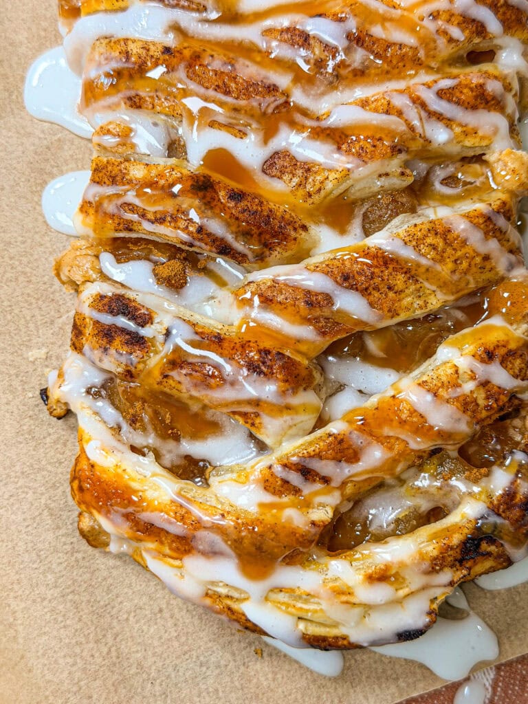 Close-up of a golden, braided pastry topped with white icing and caramel drizzle, showing a crisp texture and gooey filling, resting on parchment paper.