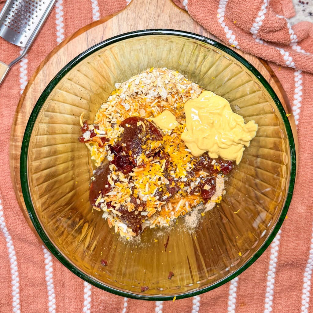 A glass bowl containing rolled oats, dried cranberries, orange zest, and a dollop of yellow mustard.