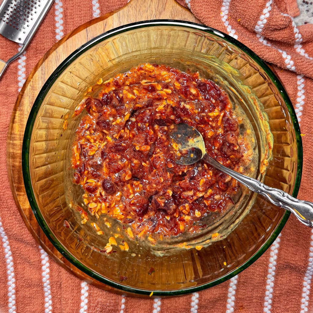 A glass bowl containing a thick, reddish-brown mixture of chopped ingredients and a metal spoon, resting on a wooden board next to a grater and a striped cloth.