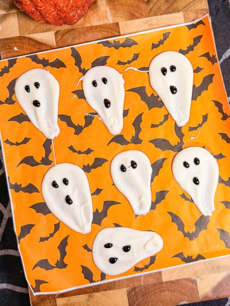 Seven ghost-shaped candies with black icing eyes and mouths, arranged on an orange sheet patterned with black bats.
