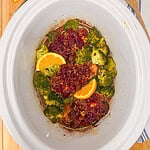 Slow cooker filled with chicken topped with cranberry sauce, orange slices, and broccoli florets.