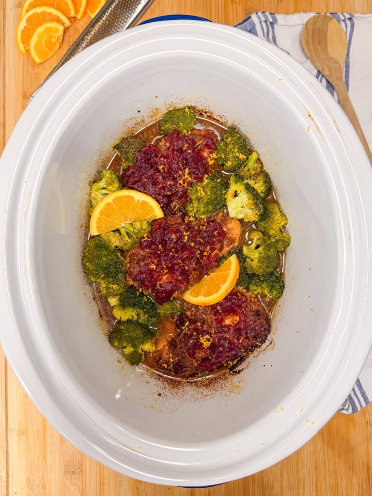 Slow cooker filled with chicken topped with cranberry sauce, orange slices, and broccoli florets.