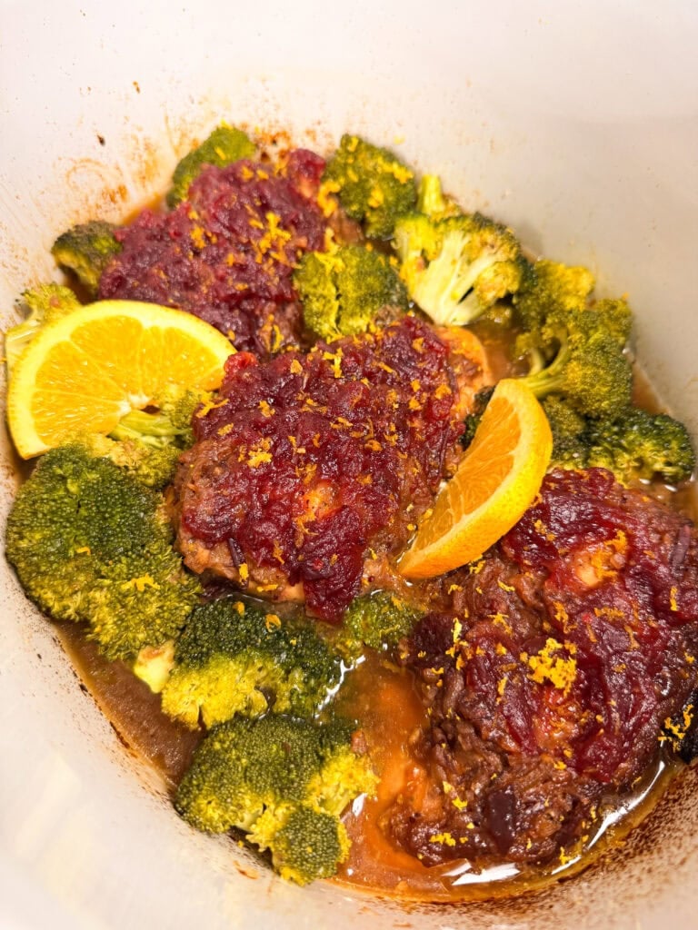 Meatballs topped with cranberry sauce and orange zest, served with broccoli florets and orange slices in a pot.