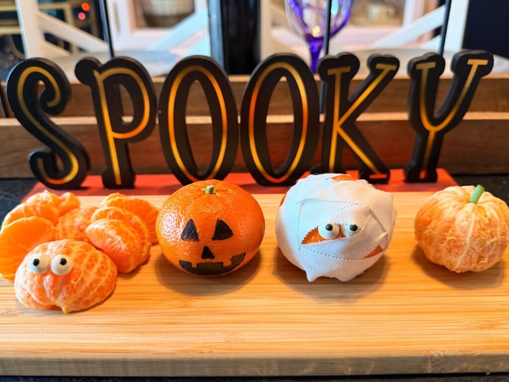 Four clementines served as easy Halloween food ideas on a wooden board: one with googly eyes, a jack-o-lantern, a gauze-wrapped mummy, and a peeled fruit, all arranged in front of a sign that reads SPOOKY.