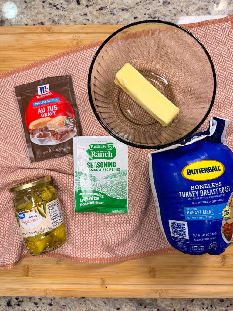 A bowl with a stick of butter, a jar of pepperoncini, ranch seasoning, gravy mix, and packaged turkey breast roast on a pink cloth on a wooden surface.