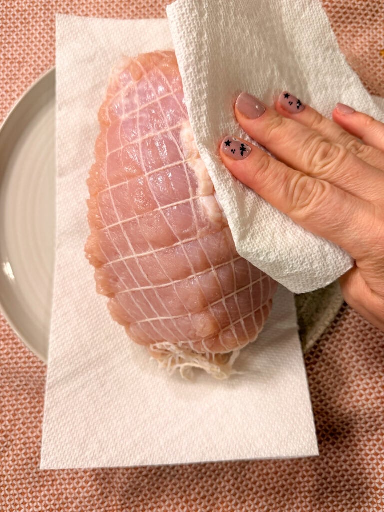A hand with painted nails pats raw, netted turkey breast dry with a paper towel on a plate.