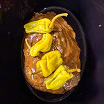 A cooked roast covered in brown gravy is topped with whole yellow pepperoncini peppers, sitting inside a black slow cooker.