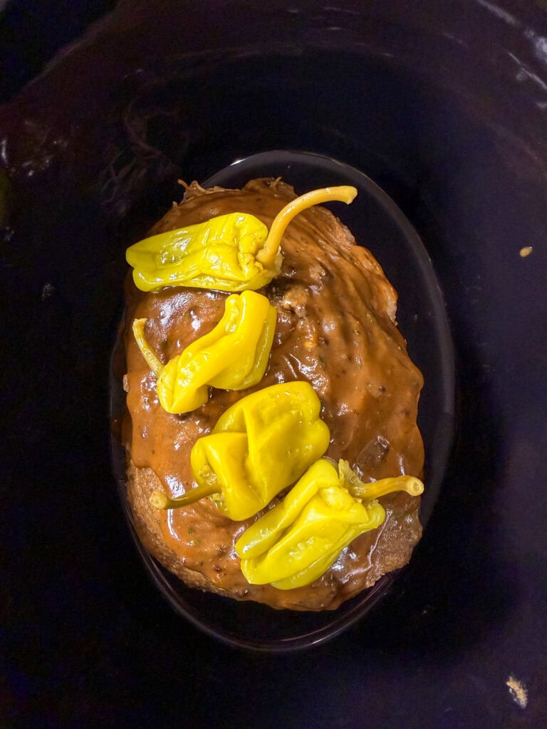 A cooked roast covered in brown gravy is topped with whole yellow pepperoncini peppers, sitting inside a black slow cooker.