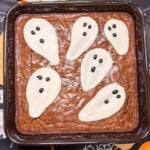 A square brownie in a baking pan topped with five ghost-shaped sugar decorations featuring dark chocolate chip eyes and mouths.