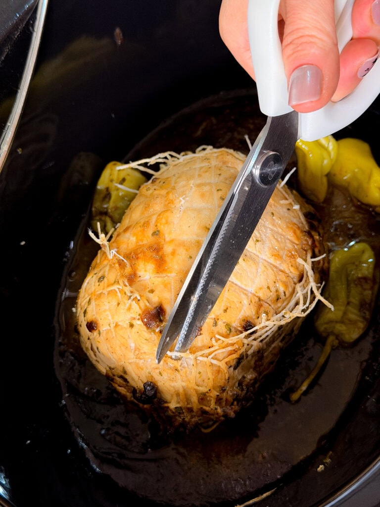 A hand uses scissors to cut string from a cooked, tied roast in a slow cooker, surrounded by juices and green peppers.