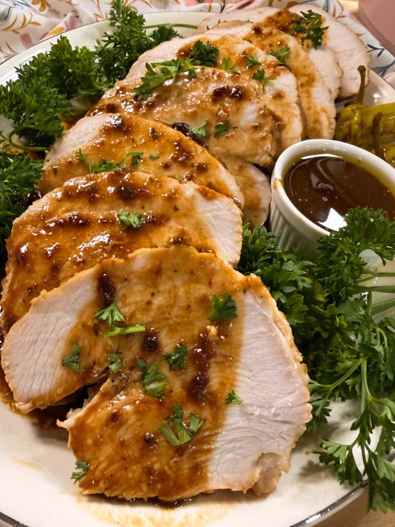 Sliced roasted turkey breast topped with brown gravy and garnished with parsley, served with extra sauce in a small cup and green herbs on a white plate.