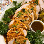 Sliced roasted turkey breast with brown gravy, garnished with fresh parsley on a white platter. A small bowl of extra sauce and parsley sprigs are beside the turkey.