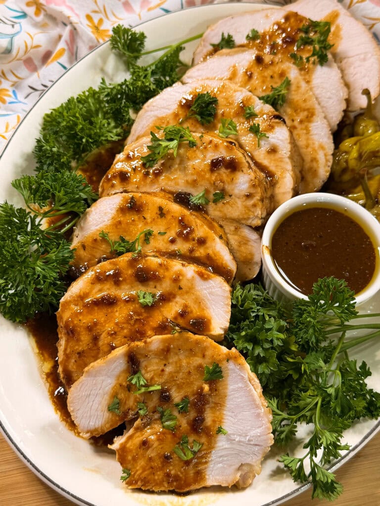 Sliced roasted turkey breast with brown gravy, garnished with fresh parsley on a white platter. A small bowl of extra sauce and parsley sprigs are beside the turkey.