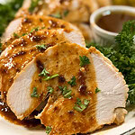 Sliced roasted chicken breast topped with brown sauce and garnished with chopped parsley, served on a plate with fresh greens and a cup of sauce in the background.