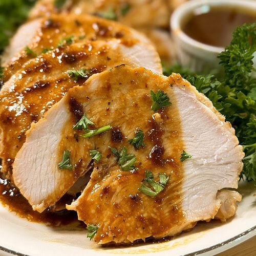 Sliced roasted chicken breast topped with brown sauce and garnished with chopped parsley, served on a plate with fresh greens and a cup of sauce in the background.