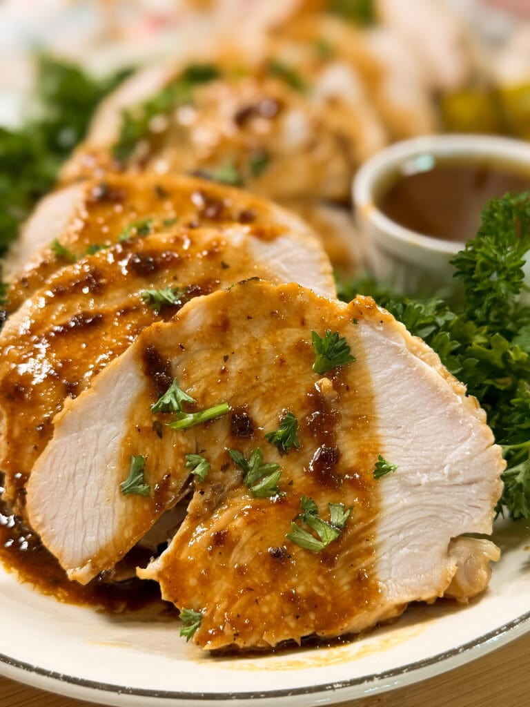 Sliced roasted chicken breast topped with brown sauce and garnished with chopped parsley, served on a plate with fresh greens and a cup of sauce in the background.