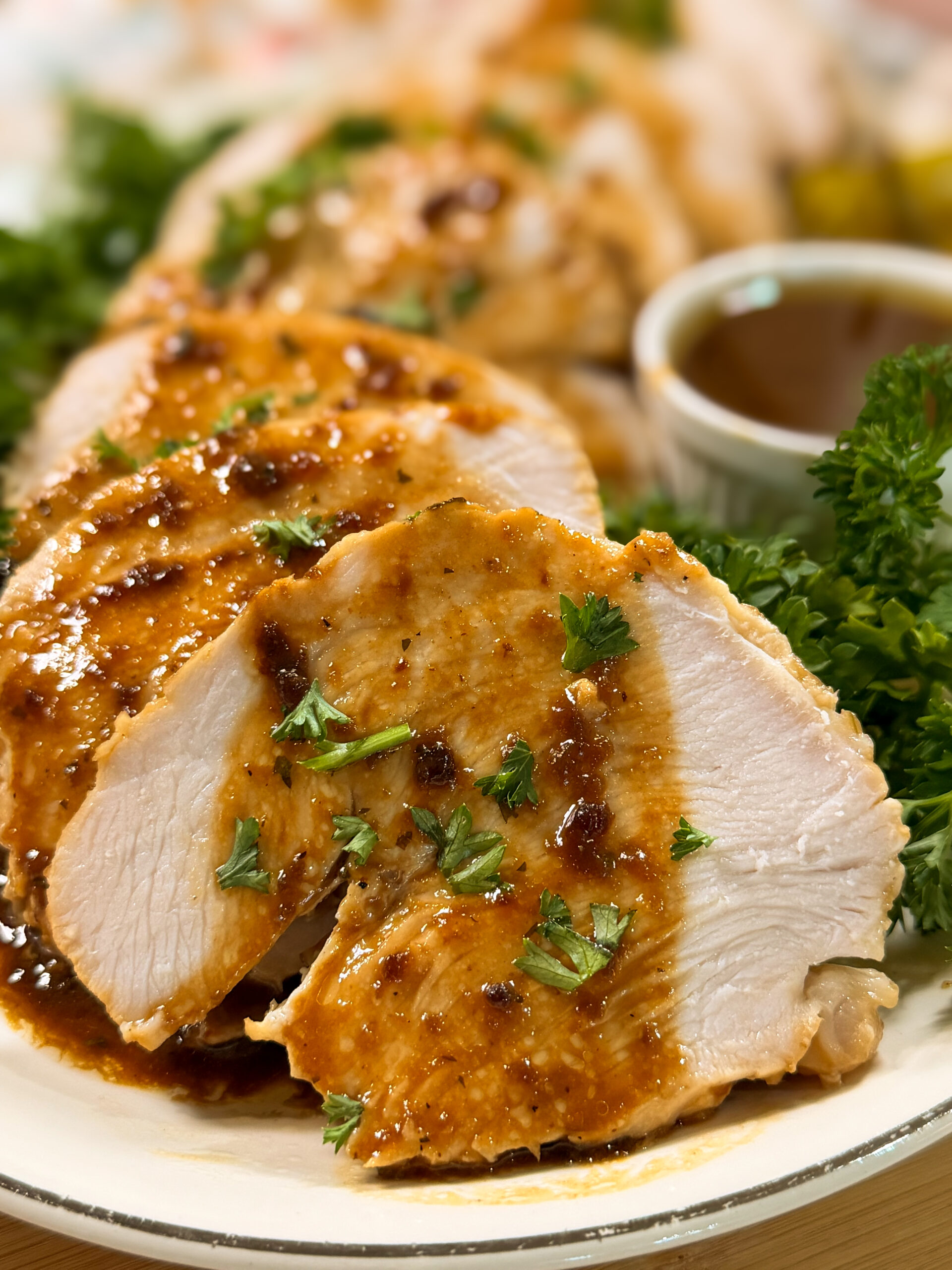 Sliced roasted chicken breast topped with brown sauce and garnished with chopped parsley, served on a plate with fresh greens and a cup of sauce in the background.