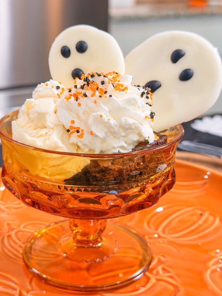 A brownie sundae with vanilla ice cream and whipped cream topped with orange and black sprinkles and two white chocolate ghost decorations in an amber glass dish.
