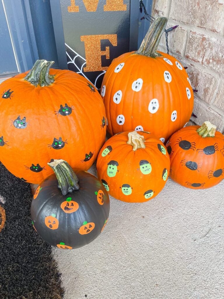 Five painted pumpkins decorated with various Halloween motifs, including black cats, white ghosts, green Frankenstein monsters, spiders, and orange jack-o-lantern faces.