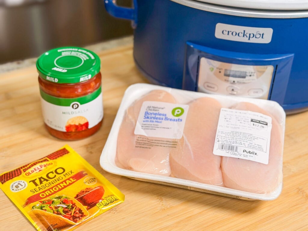 A blue Crock-Pot sits behind a jar of mild salsa, a package of raw boneless skinless chicken breasts, and a packet of Old El Paso taco seasoning on a wooden surface.