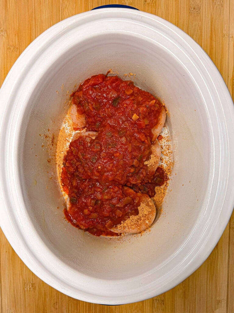 Raw chicken breasts topped with salsa and dry seasoning in a slow cooker.