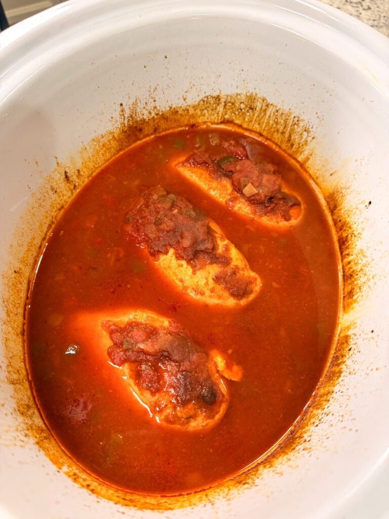 Three chicken breasts simmering in a red tomato-based sauce inside a slow cooker.