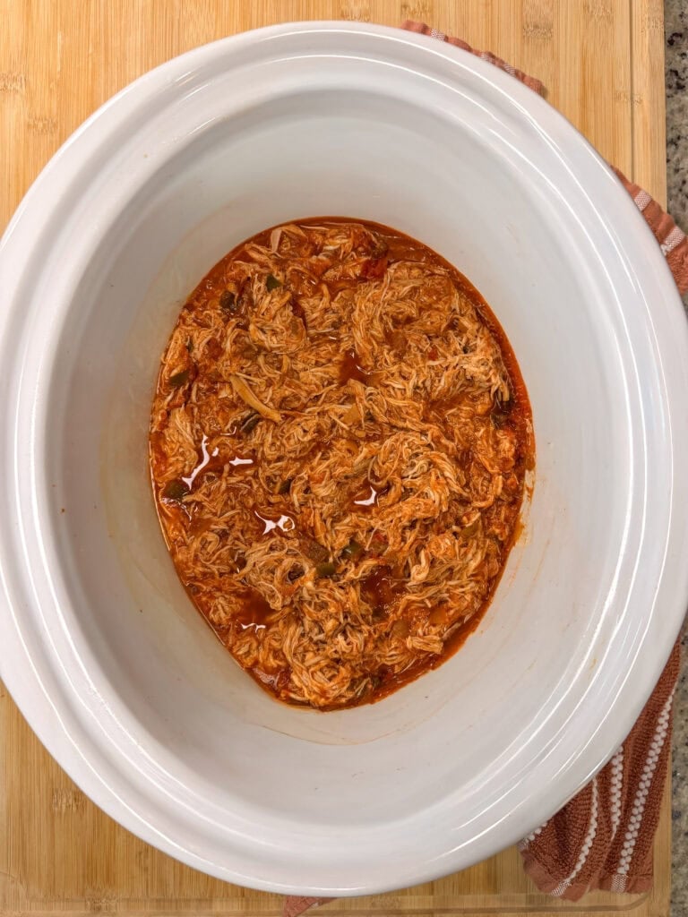 Shredded chicken in a tomato-based sauce inside a white slow cooker crock.