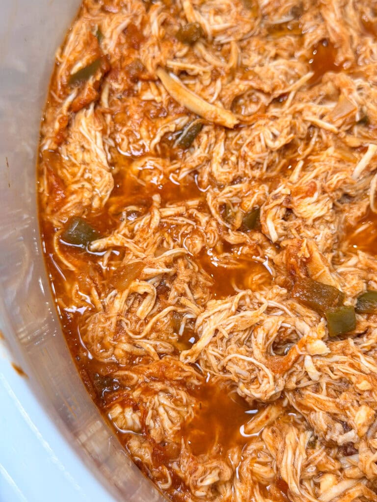 Shredded chicken in a red sauce with pieces of green pepper in a slow cooker.