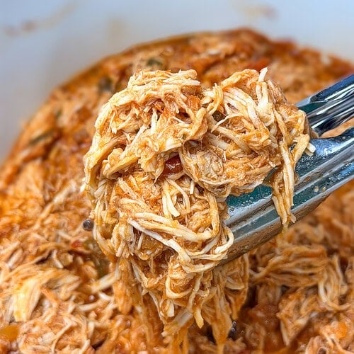 Metal tongs lifting a portion of shredded chicken coated in a red tomato-based sauce.