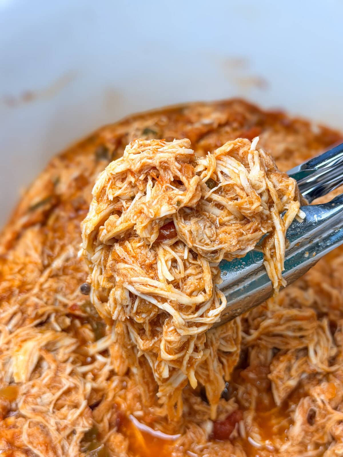 Metal tongs lifting a portion of shredded chicken coated in a red tomato-based sauce.