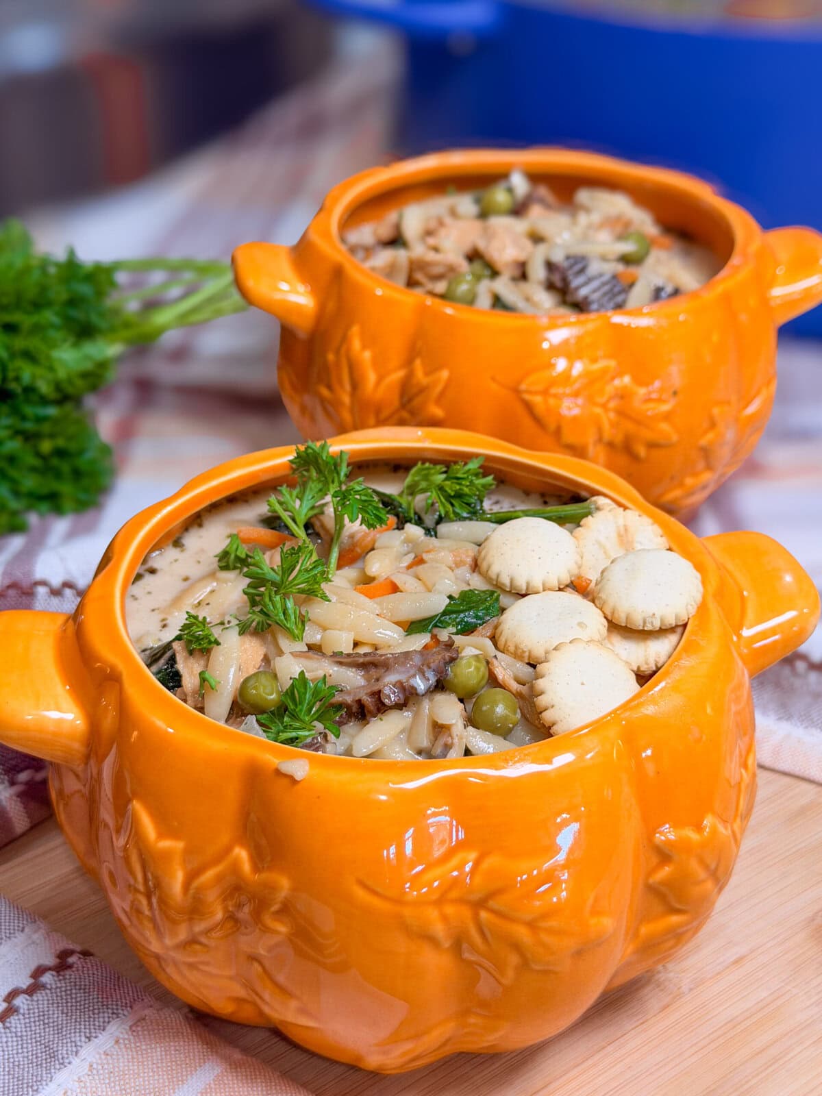 Two orange pumpkin-shaped bowls filled with chicken and noodle soup topped with crackers and parsley.