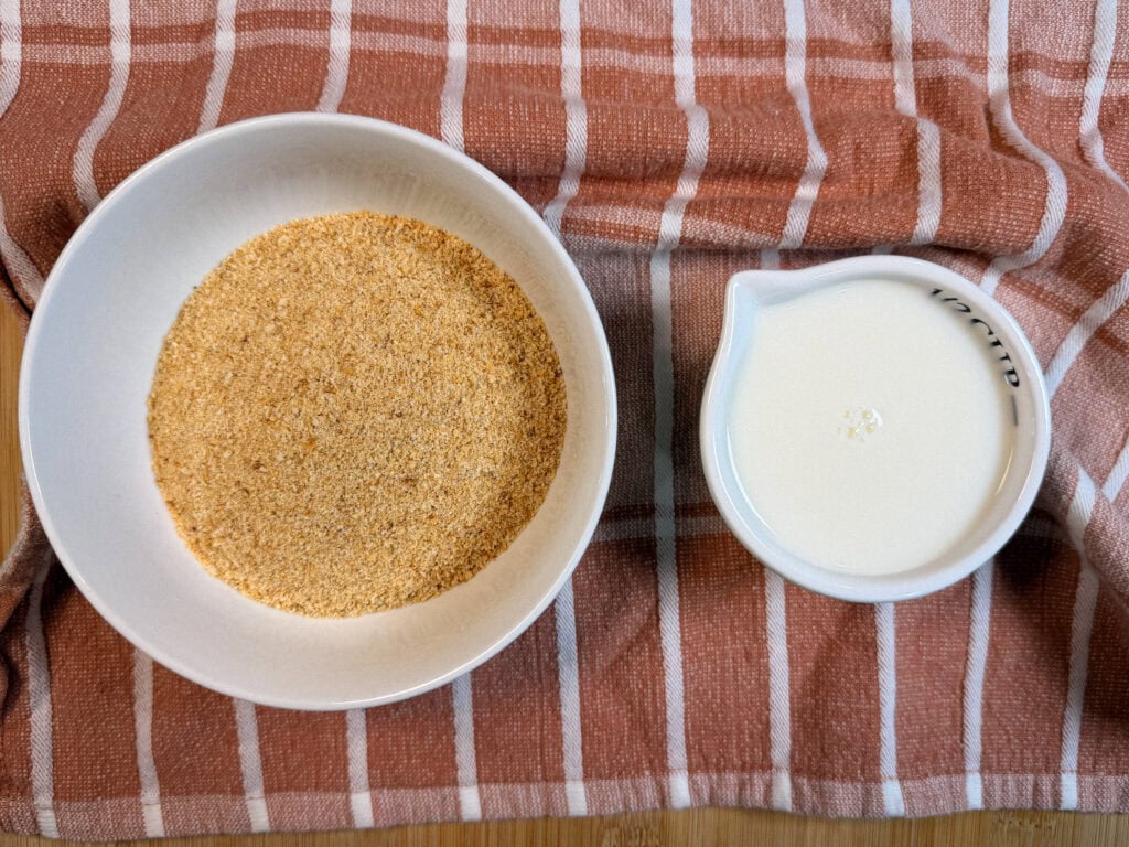A bowl of breadcrumbs and a small pitcher of milk on a plaid cloth.
