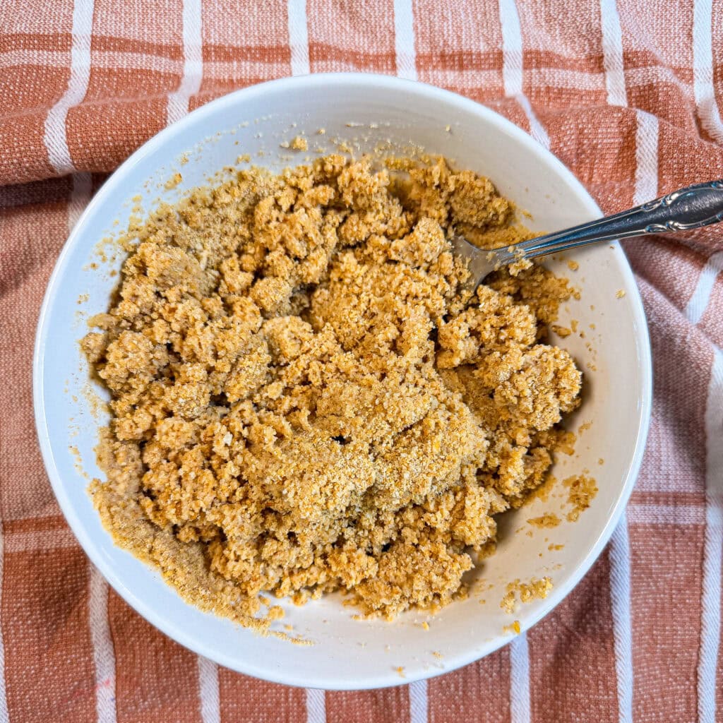 A white bowl filled with a coarse, golden-brown crumb mixture and a metal spoon, sitting on a striped cloth.