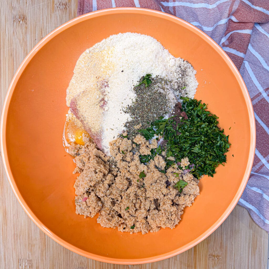 An orange mixing bowl containing an egg, breadcrumbs, chopped fresh herbs, and various seasonings.