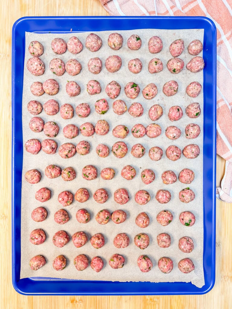 A blue baking sheet lined with parchment paper holds rows of raw meatballs.