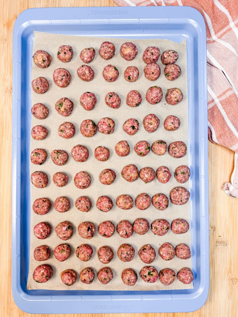 A light blue baking sheet lined with parchment paper, filled with rows of raw, round meatballs.