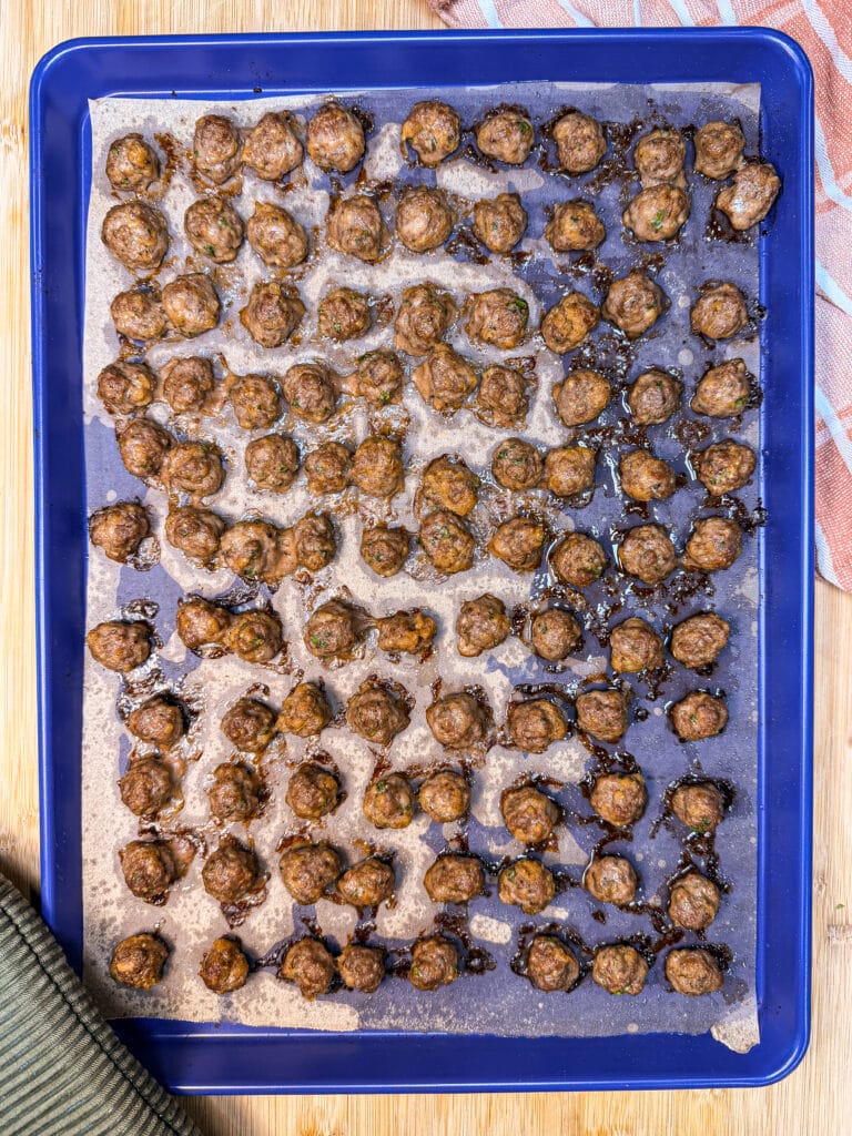 A large blue baking sheet lined with parchment paper covered in many small, freshly baked meatballs.