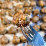 A single cooked meatball held on a silver fork, with many more meatballs visible in the blurred background.