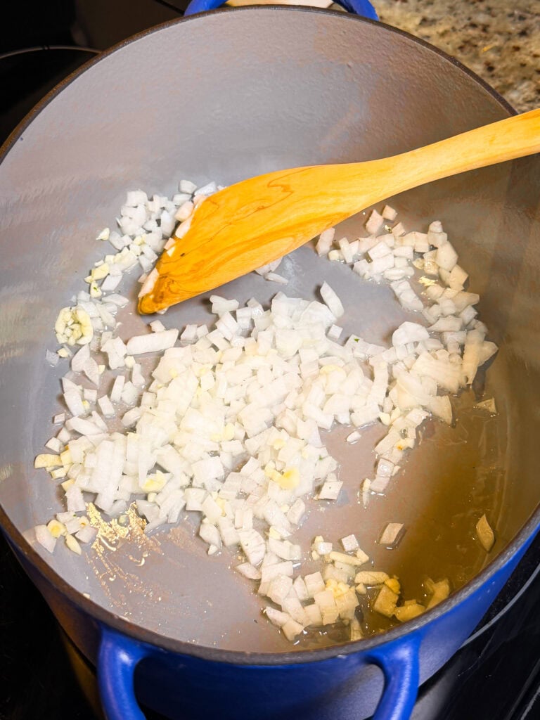 Diced onions and minced garlic cooking in a blue Dutch oven with a wooden spatula.