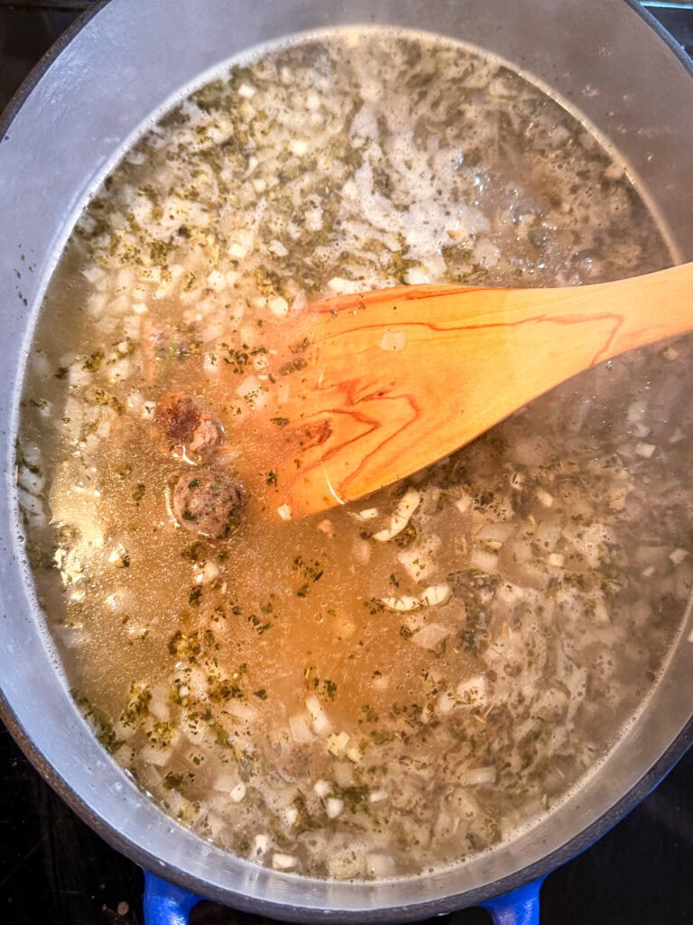 A wooden spoon stirs a pot of simmering soup containing minced onions, herbs, and small meatballs.