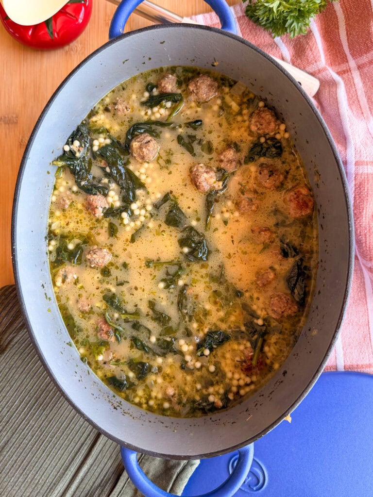 A blue Dutch oven filled with Italian wedding soup containing meatballs, spinach, and acini de pepe pasta.