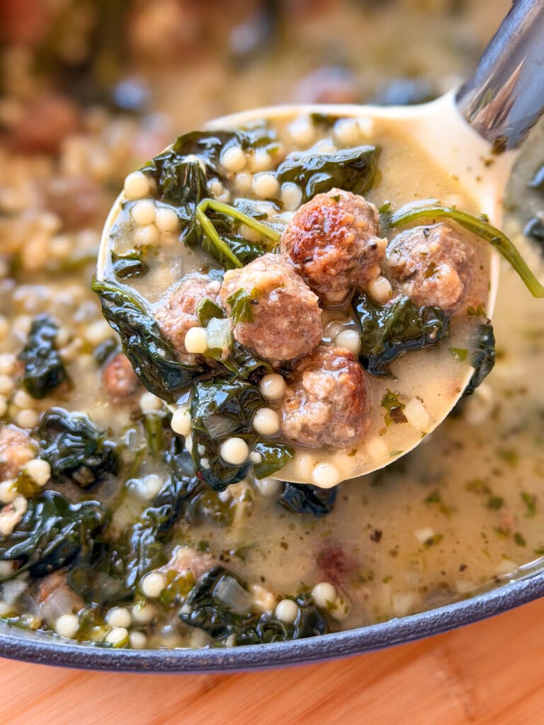 A spoonful of Italian wedding soup featuring meatballs, small pasta pearls, and greens held over a pot.