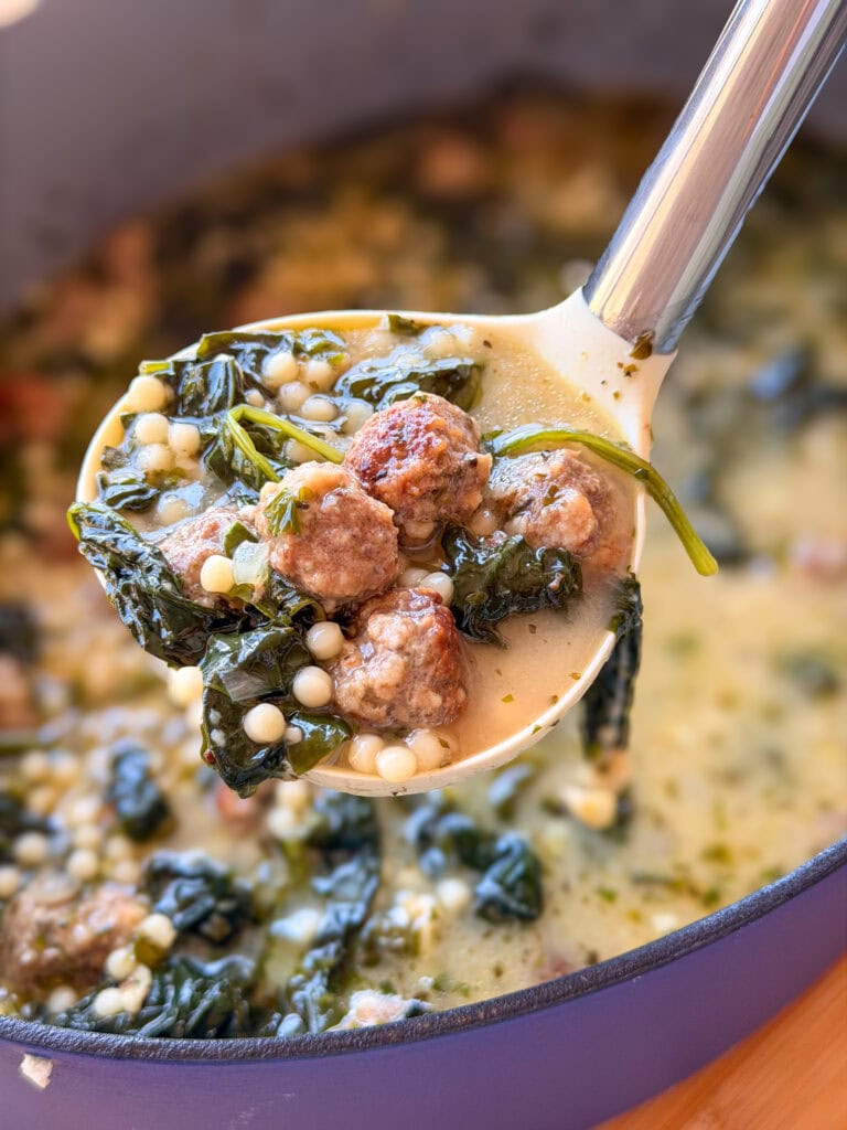 A ladle lifting wedding soup containing meatballs, leafy greens, and tiny pasta beads from a pot.