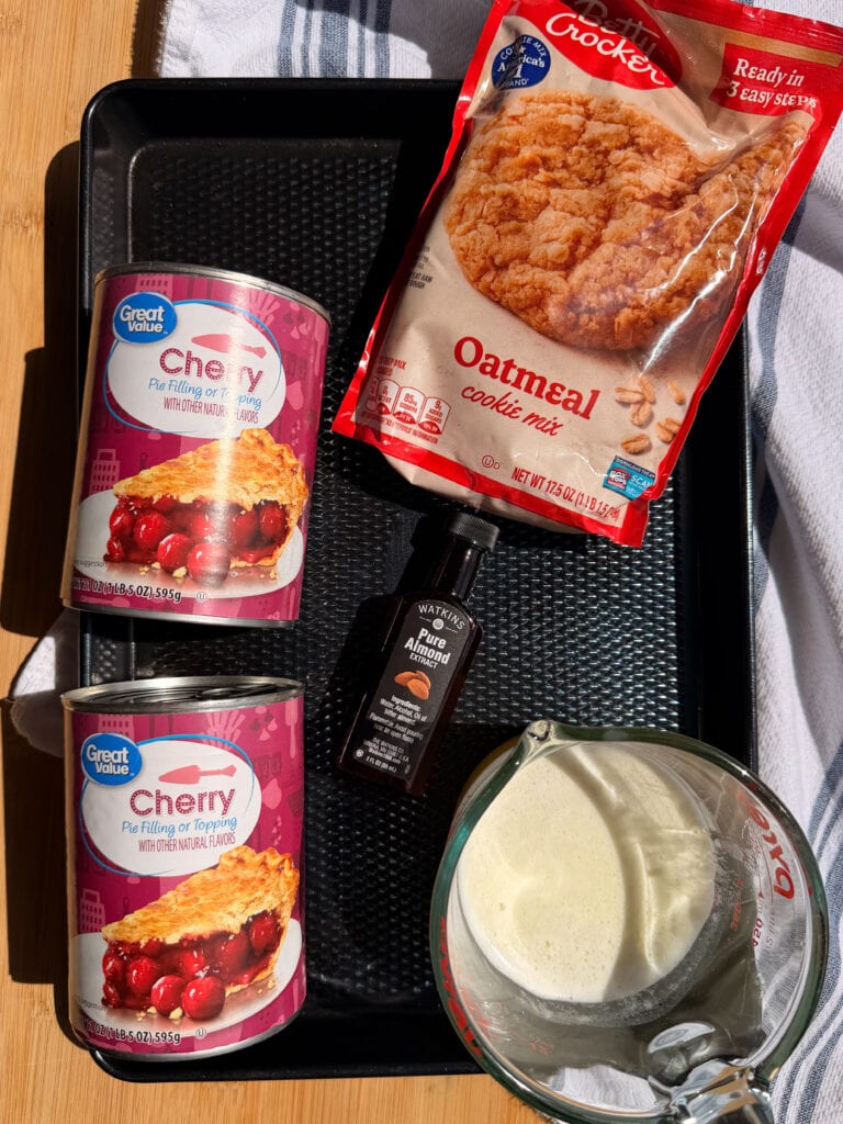 Two cans of Great Value cherry pie filling, a bag of Betty Crocker oatmeal cookie mix, a bottle of Watkins pure almond extract, and a measuring cup of milk on a black tray.