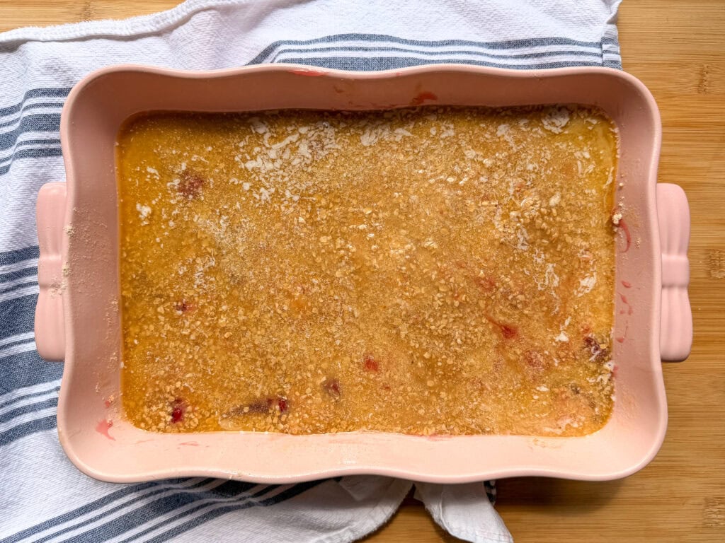 A rectangular pink baking dish containing an unbaked fruit crumble with a golden, oat-based topping on a striped kitchen towel.