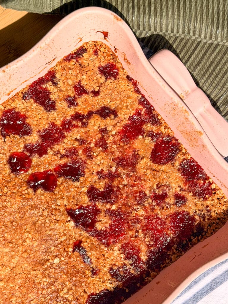 A fruit crumble with a golden-brown oat topping and patches of red berry filling in a rectangular pink baking dish.