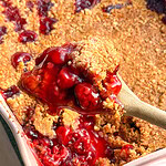 A wooden spoon lifts a scoop of cherry crisp from a pink baking dish filled with red fruit filling and a golden brown crumb topping.